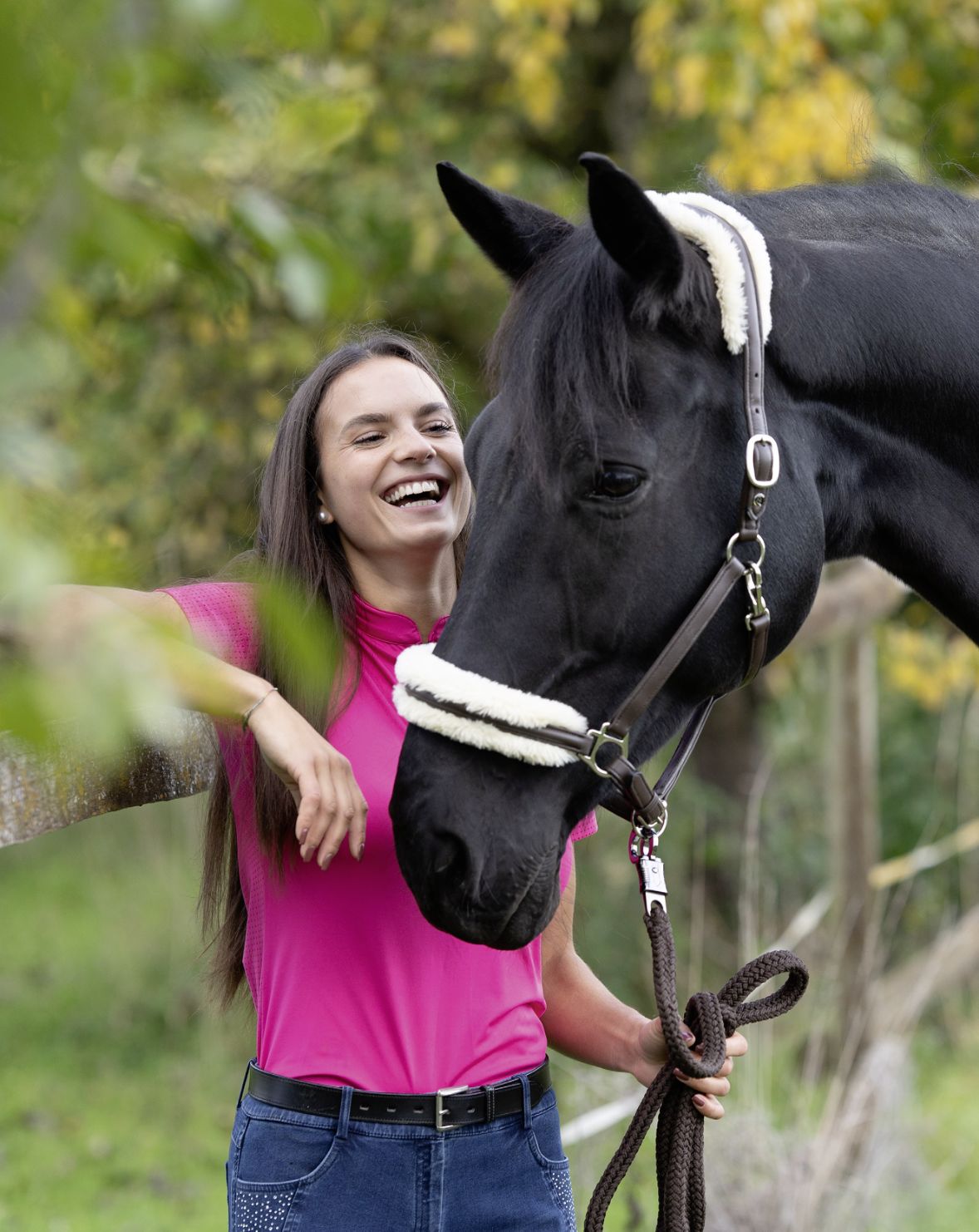 Eine lachende Frau in pinkem Oberteil streichelt ein schwarzes Pferd mit Halfter im Freien.