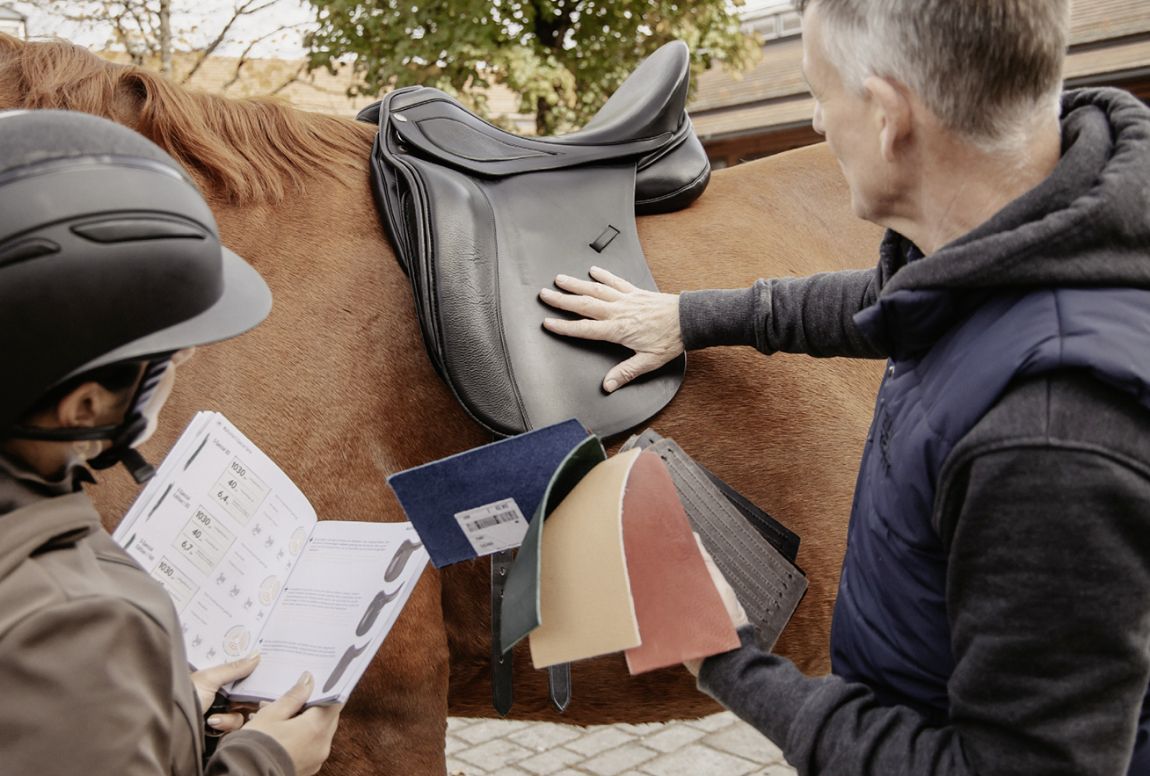 Ein Reiter in Helm und Jacke betrachtet ein Sattelkatalog, während ein Sattler verschiedene Sattelmaterialien an einem Pferd demonstriert.